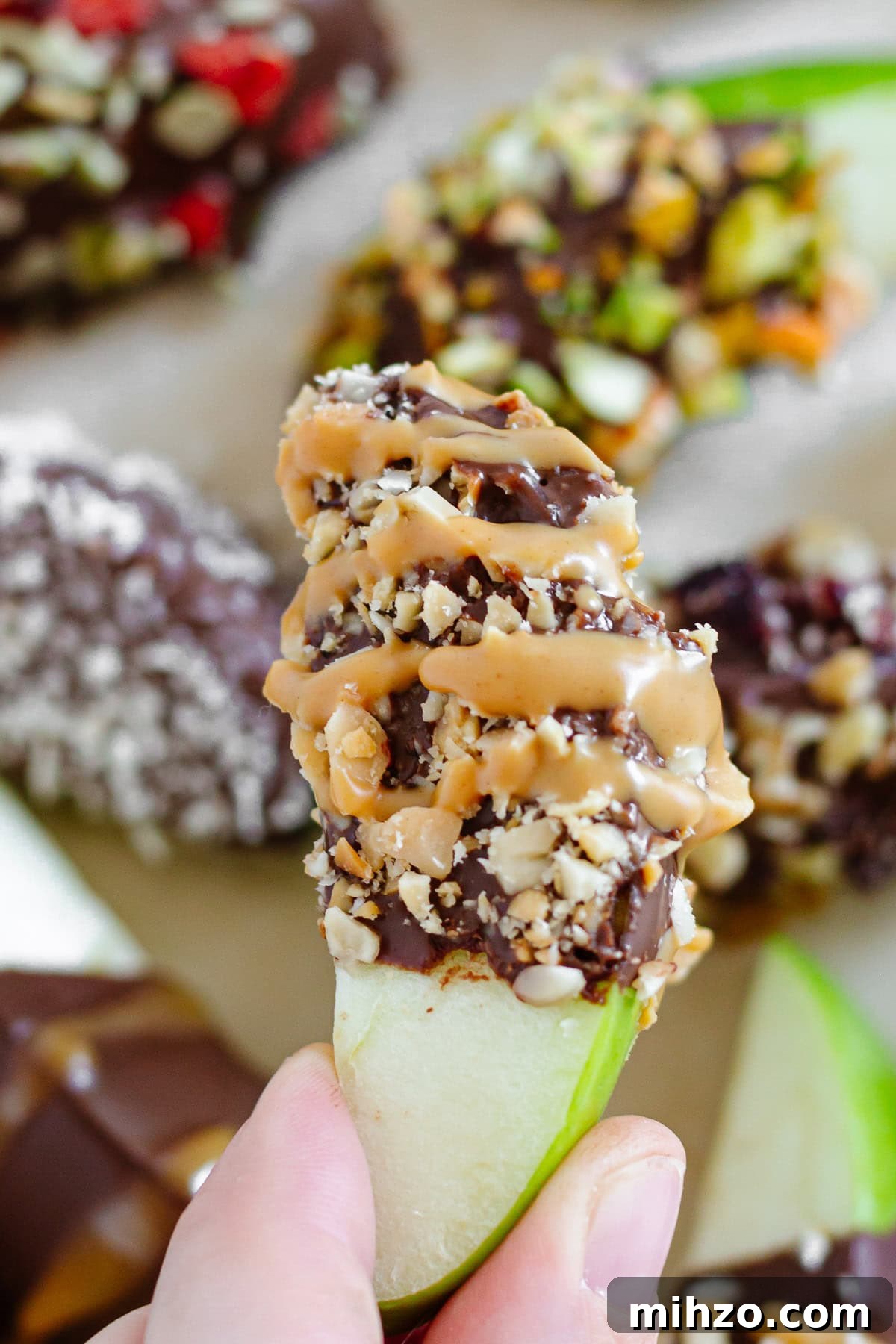 Chocolate-Dipped Apple Slices for Snacks & Parties 11 Hand holding up a chocolate covered apple slice topped with crushed peanuts and peanut butter drizzle.