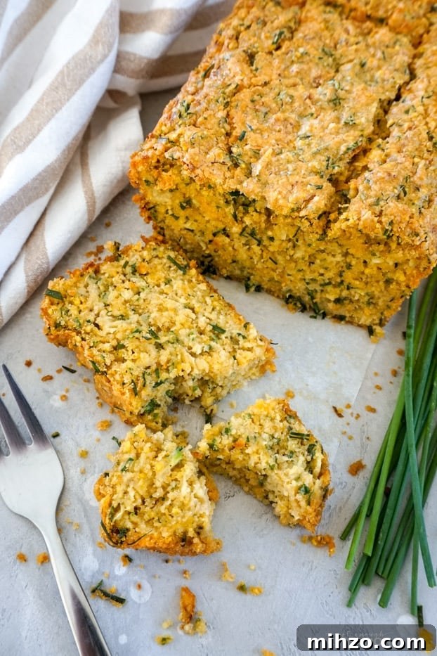 A slice of cheddar chive zucchini bread broken in pieces, next to a fork, with the bread loaf in the background and fresh chives on the side.