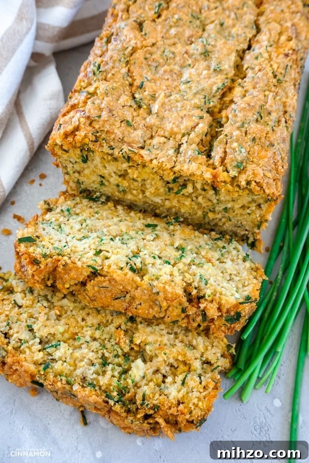 A loaf of cheddar chive zucchini bread, cut partially in two slices, with a few chive sprigs on the side and a striped brown and white napkin 