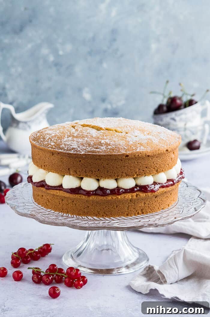 Vegan Victoria sponge cake on a cake stand. Vegan victoria sponge cake on a glass cake stand with fruit and crockery in the background.