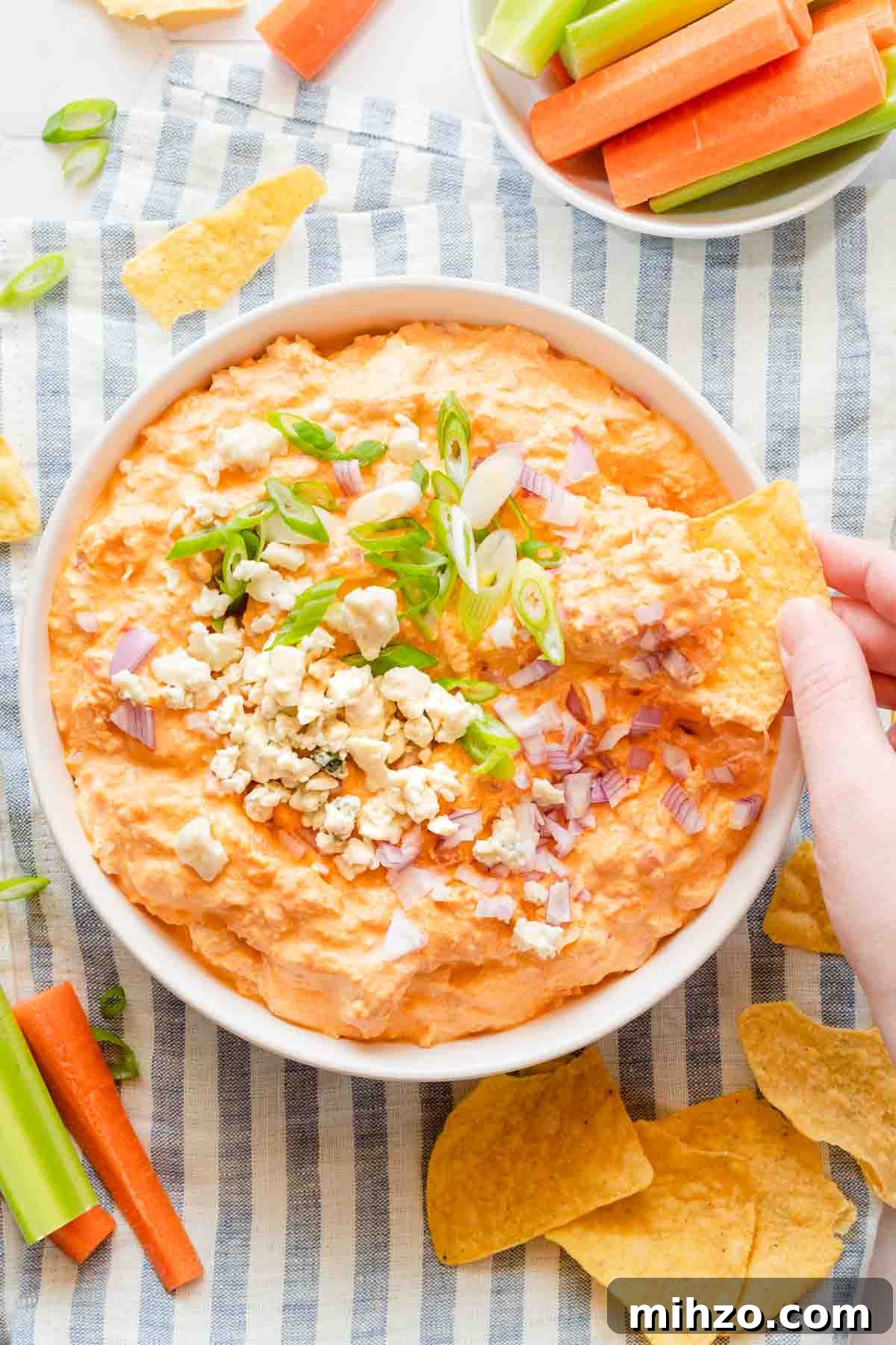 hand scooping chip into bowl of buffalo chicken dip with veggies and chips on the side.