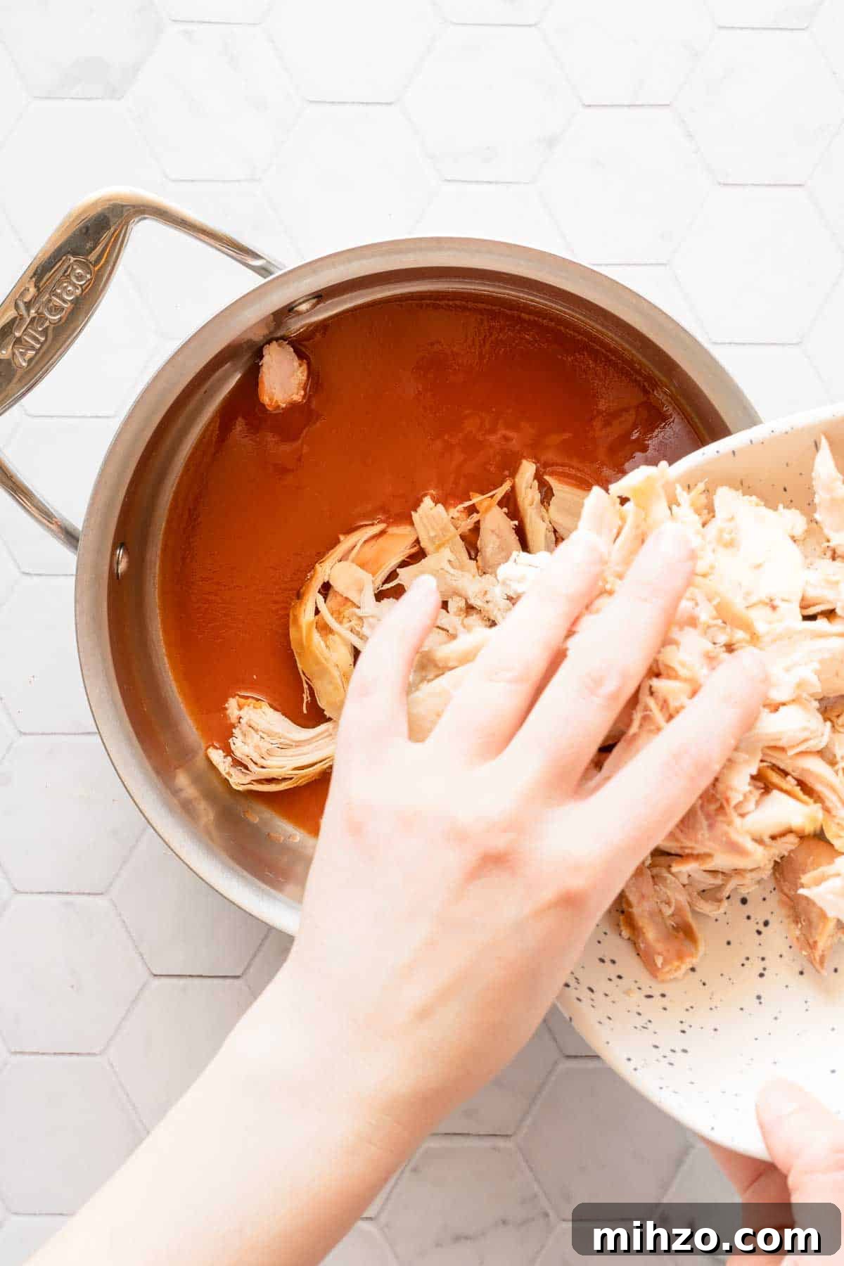 shredded chicken being added into saucepan with hot sauce inside.
