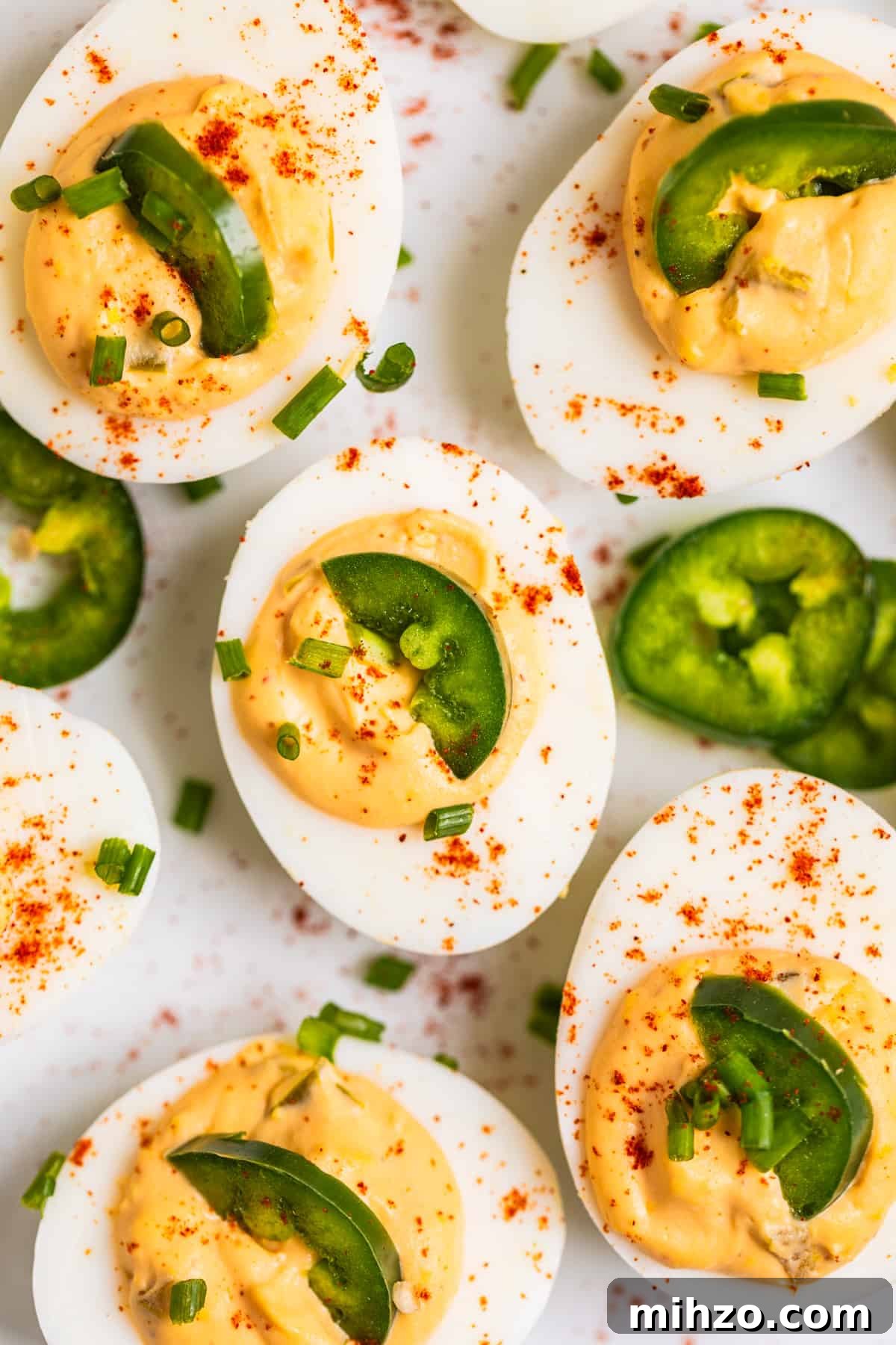 Spicy Deviled Eggs with Sriracha and Smoked Paprika 2 White dish with spicy deviled eggs topped with half slice of jalapeño and a sprinkle of paprika and chives.