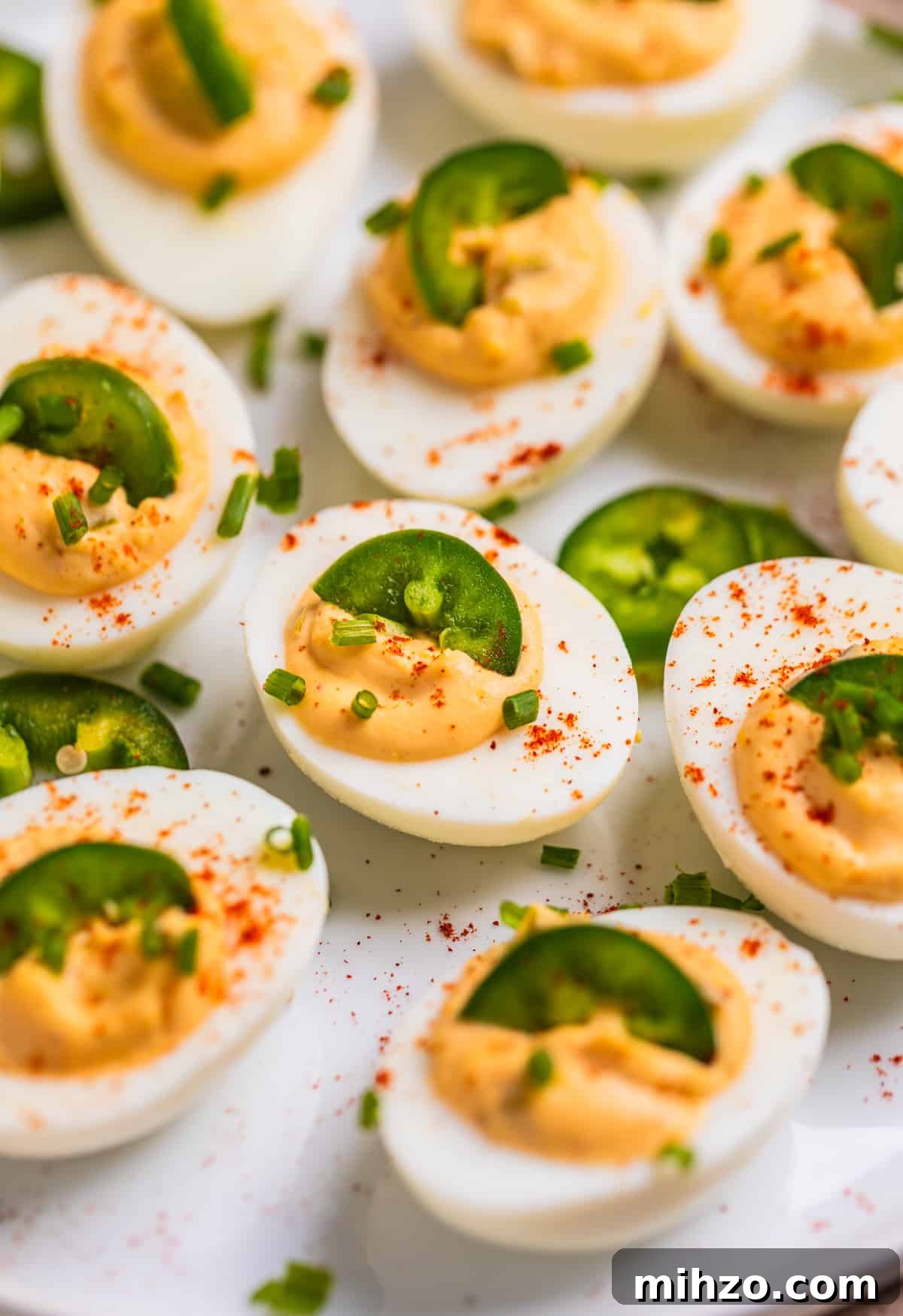 Spicy Deviled Eggs with Sriracha and Smoked Paprika 10 White dish with spicy jalapeño deviled eggs garnished with chives, paprika and sliced jalapeño.